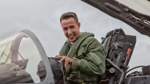 A pilot in a green flying suits smiles as he sits in the cockpit of an RAF jet, with the canopy raised above his head.