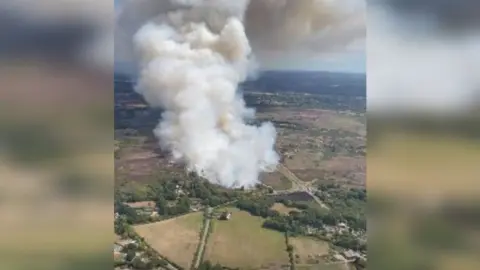 Aerial view of white smoke plume coming from heath fire surrounded by patchwork of green fields.