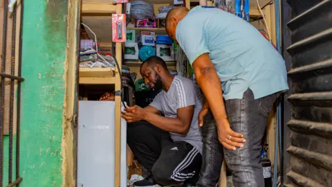 University of Hertfordshire Two men in a small room, looking at a power box. One is kneeling and the other is bending over. There are items on shelves behind them.