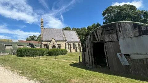 A small stone church with green hedges to the front stands among farm buildings, some in a dilapidated condition, on a bright sunny day.