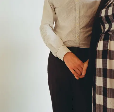 Gemma Briggs The middle portion of a woman standing up wearing trousers and a shirt. Her right hand appears to be holding another person's hand in front of her crotch, although the other person is hidden behind some patterned material. 