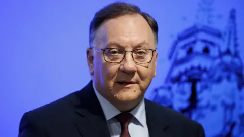PA Media Rev Trevor Gribben, pictured from the chest up. He is wearing a black suit, blue shirt and dark red tie. He has short brown hair. He is standing in front of a blue background. 