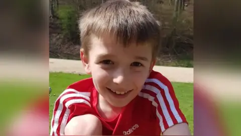 A young boy with short brown hair smiling at the camera. He's wearing a red and white Adidas shirt and is in a garden, with grass and trees behind him