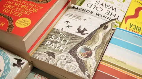 Getty Images A book cover, entitled the Salt Path, with artwork depicting a coastal scene. It is placed on a table stacked with other books around it.