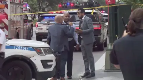 Police are standing outside the site of the Manhattan shooting, surrounded by police tape, flashing lights and vehicles.