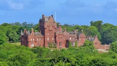 Anne Burgess A red stone castle set among plentiful green trees