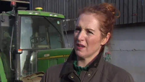 Clare Wise, a woman of about 40 with auburn hair tied back, is standing in front of a tractor.