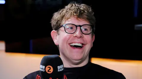 Comedian Josh Widdicombe laughs while sat down in a BBC Radio Two studio. He is wearing black-rimmed classes and a black top. He is sat in front of a microphone with BBC Radio Two's logo on it.