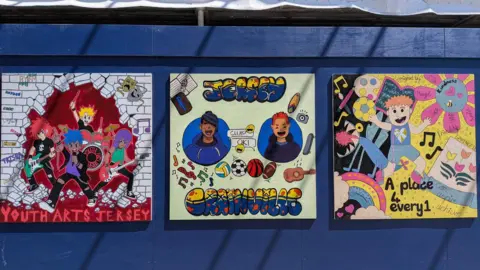 A selection of artwork displayed on a hoarding outside a construction site. The art on the left shows a band and says 'Youth Arts Jersey'. The second painting shows two smiling characters and says 'Jersey Grainville'. The third is mostly yellow and pink and hows a boy playing with a ball. Words over the top read 'A place 4 every1'.