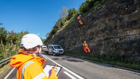 A person in a high vis jacket holding a clipboard and wearing a white helmet looks at a rock face. On the rock face are two people in full orange high vis abseiling. On the road below is a parked white van.