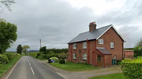 A long straight road with green bushes and trees on either side, and a large red brick house on the right hand side. The house has a path leading to a driveway and has white windows and red drainpipes