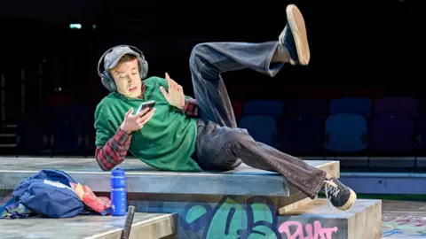 Dan Rainford in Top G's Like Me. The actor, wearing a green baggy polo and blue jeans is laying on his side in a skatepark and scrolling on his phone as he listens to content through headphones.