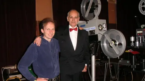 John Clancy John Clancy and Keith Wilton posing next to a large film projector. John is wearing a blue shirt and Keith is wearing a tuxedo with a red tie.