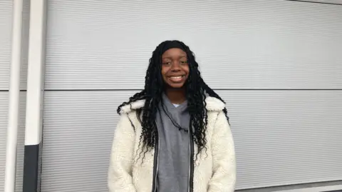 Miracle Moyo stands against a grey background, she is smiling and wears her long black hair in plaits held back with a black hair band. A grey hoodie is visible under her cream coloured woolly fleece jacket.
