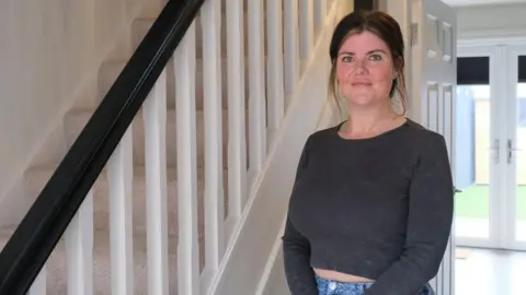 Prima Rachael McCoy, standing inside the hallway of her home. She has brown shoulder length hair in a ponytail, and is wearing a blue-grey top.