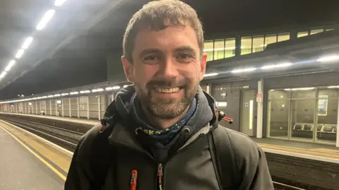 BBC A man in a black coat stands on a train platform. He is wearing a hoody. 