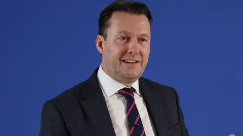 Russell Findlay has short dark hair and wears a dark suit and white shirt with a navy and red striped tie. He is mid speech and in front of a blue background