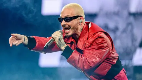 J Balvin performing in a red jumpsuit, he is pointing at the crowd.