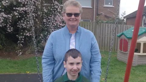 Lesley has grey short hair, sunglasses and a blue knitted cardigan on. She is standing in front of a tree with pink flowers, a green, red and cream wendy house and she has her hands on her son's shoulders. He is sitting on a swing, he has dark short hair and is wearing a green school jumper. He's smiling at the camera. 