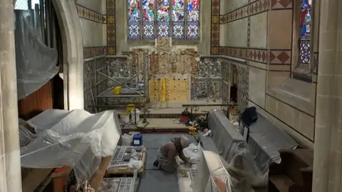 Jamie Niblock/BBC Wall bare of mosaic exposes brick and wood panelling. A woman is kneeling on the floor sorting through stones. The church pews are covered with protective plastic sheeting. Other examples of mosaic wall-coverings can be seen on two walls in the altar area of the church. A large multi-coloured stained-glass window is above the chancel.