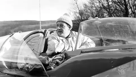 Tom Buist/Mirrorpix/Getty Images A close-up black-and-white photograph of Donald Campbell in the cockpit of Bluebird. He is wearing a woolly hat and a zipped top. The craft canopy is open. Trees and hills can be seen in the background.