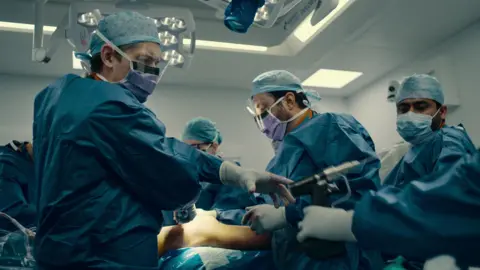 Doctors and nurses wearing blue scrubs and facemasks and gloves. They are carrying out surgery on a patient.
