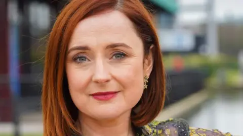 Siobhan O'Neill has short red hair. She is wearing a green sequin top and has star-shaped earrings. The background is blurred behind her.