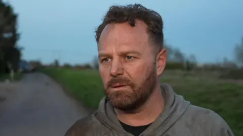 Qays Najm/BBC Billy Cunningham has short brown hair and a brown beard. He is wearing a black T-shirt and grey hoodie and is standing in front of a river bank which is on the right.