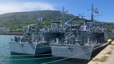 A warship on the water docked with a union jack visible and a Ukraine Navy flag also visible with some green hills in the background