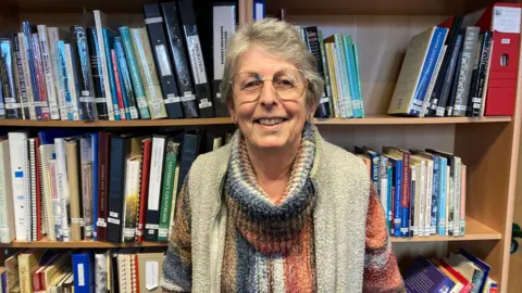 Lyn Bidgood is standing in front of a book shelf. She is wearing a knitted jumper and a fleece vest. She is wearing glasses and is smiling at the camera.