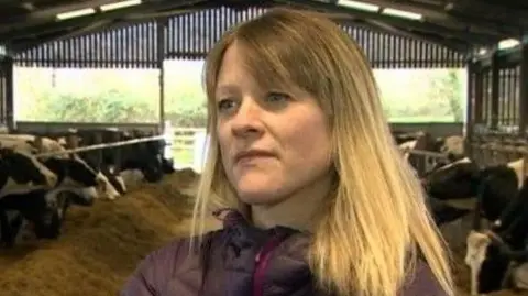 Gwen Rees Gwen Rees has long blond hair and is wearing a purple coat. She is standing in a shed in which several cows are feeding