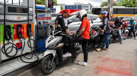 A worker helps a motorcyclist top up fuel for his bike. Behind him are more than five other bikers queuing for fuel.