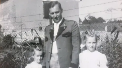 A black and white image of a man in a military-style jacket holding the hands of his two daughters in white blouses.