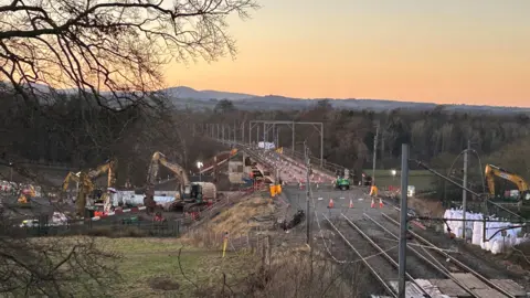 Yellow cranes and excavators have surrounded the concrete Clifton rail bridge. Dozens of white and orange traffic cones are lined on the structure as the sunset turns the sky behind pink.