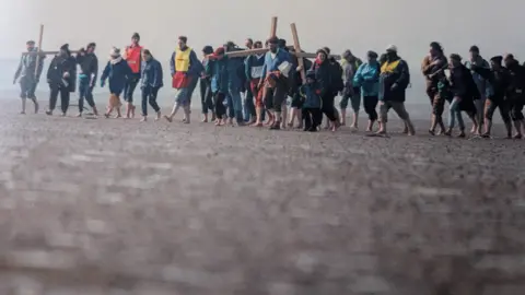 Jacqui Feline A snapshot of about 30 people all barefoot carrying crosses with their trouser legs rolled up walking on wet dark sand 