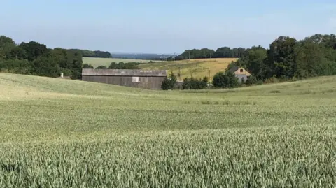 An image showing a field sloping down towards a grey farm building with further hills and trees in the distance beneath a blue sky.