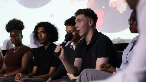 ISCRE A young man holds a microphone and talks while others on the stage look on. He is wearing a black polo shirt. 