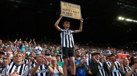 A boy is holding up a sign asking for a shirt in a sea of Newcastle supporters 