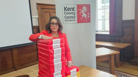 Kent County Council leader Linden Kemkaran, wearing a red jacket, leans on a large pile of packets of A4 paper piled on a table at County Hall in Maidstone, each one labelled with the number 1,000.