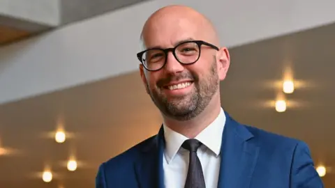 Ben Macpherson, who is bald with glasses, wearing a blue business suit 