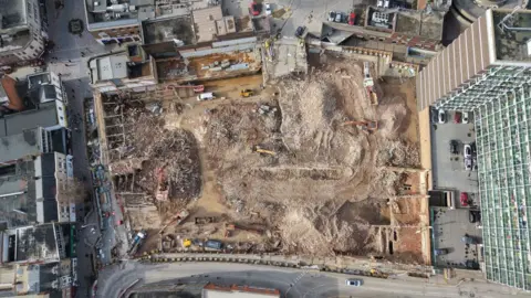 An aerial shot  from directly above the site shows buildings fully demolished with some debris and mounds of dirt. There are also construction vehicles. 