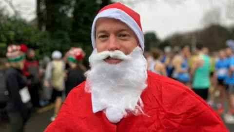 Man dressed as Santa with fake white beard stood in front of runners.