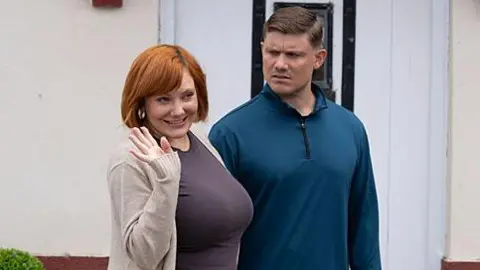 BBC/HOT OLIVES PRODUCTIONS LIMITED Sophie Willan, who has red mid-length hair styled into a loose bob, smiles as she waves while standing next to man outside a front door.