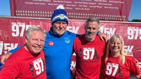 Erica Dillon Group picture of Sammy, Steve, Jamie and Margaret with their arms around each other and smiling. Steve is wearing his blue Everton fleece with the other three wearing red 97 tops. they are in front of branding saying "Run for the 97" and a big red sign above the names of the 97 victims.