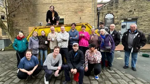 People's Property Portfolio A group of people gathered outdoors on a cobblestone street in front of a large yellow skip placed against a tall brick wall. One person is sitting on top of the skip, while the rest stand or crouch in front of it. The surrounding area features old stone buildings with arched windows and doors, and a leafless tree on the left side.