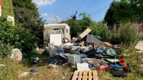 Piles of discarded furniture and rubbish on private land next to the roadside at Baguley Crescent. There is a derelict caravan behind the rubbish pile, with security fencing and overgrown plants visible.