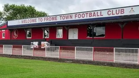 A photo of Badgers Hill stadium, the grass pitch and goal in front of a covered terrace with the words 'Welcome to Frome Town Football Club'
