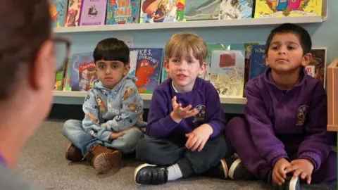 Three reception age children sit on the carpet in a classroom facing a teacher who is sitting on the floor opposite them, behind them is a row of children's books