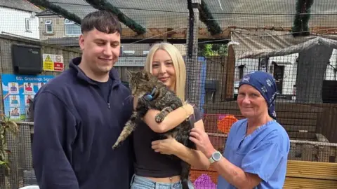 A man, and two women pose holding a large tabby cat