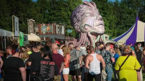 A crowd of people outdoors with the tops of tents and a large painted sculpture of a man's head in the background and a row of trees behind that
